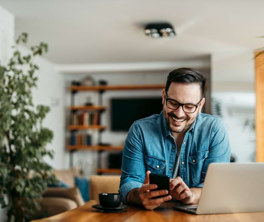 Usměvavý muž s brýlemi používá smartphone při práci na notebooku z domu.